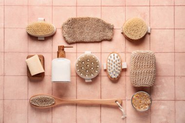 Farklı masaj vücut fırçaları, sabun çubuğu ve pembe karo arka planda bir şişe kozmetik ürünü içeren kompozisyon