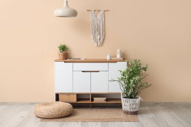 Oturma odasının iç tarafındaki bej duvarın yanındaki çekmeceler, puf ve bitkiler.