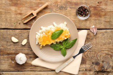 Ahşap zemin üzerinde fırında makarna, sarımsak ve fesleğen yapraklarıyla dolu bir tabak.