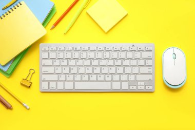 Sarı zemin üzerinde modern bilgisayar klavyesi, fare ve kırtasiye malzemesi