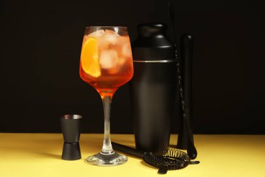 Barmen seti ve bir bardak Aperol sprey kokteyli siyah duvarın yanındaki sarı masada.