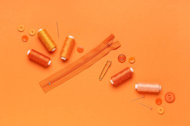 Top iğneli iplik makaraları, düğmeler ve turuncu arka planda fermuar.