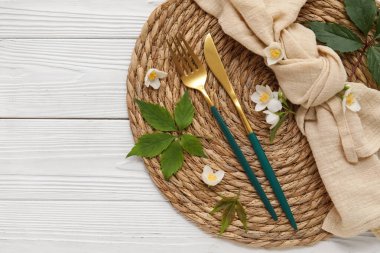 Çatal bıçaklı hasır, peçete, yapraklar ve yasemin çiçekleri beyaz ahşap arka planda