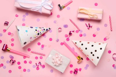 Hediye kutuları, konfetiler, düdükler, mumlar ve pembe arka planda yılan yağı olan koni parti şapkaları.