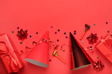 Kırmızı arka planda düdüklü koni parti şapkaları, hediye kutuları, yılan yağı ve konfeti.