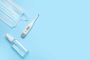 Tıbbi maskeli elektronik termometre ve mavi arka planda bir şişe antiseptik.