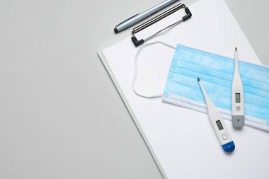 Gri arka planda panosu ve tıbbi maskesi olan farklı elektronik termometreler.