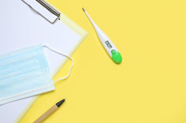 Sarı arka planda pano, kalem ve tıbbi maskeli elektronik termometre.