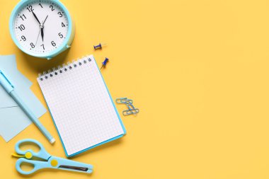 Sarı zemin üzerinde kalem, ataç, alarm saati, yapışkan notlar ve makas bulunan defter