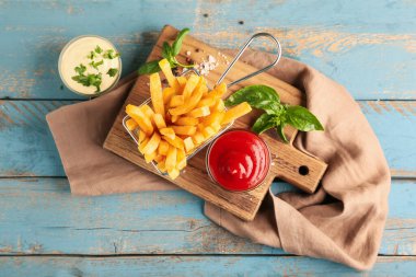 Deep fryer basket with tasty french fries and bowls of sauces on wooden background