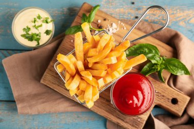 Deep fryer basket with tasty french fries and bowls of sauces on wooden background