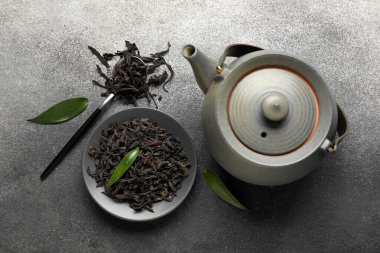 Kuru siyah çay yapraklı tabak, gri grunge arka planda kaşık ve çaydanlık.