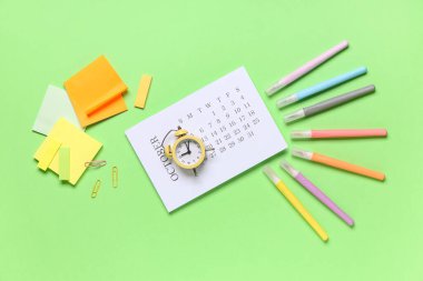 Alarmlı saat, yapışkan notlar, çıkartmalar, ataçlar ve yeşil arkaplandaki keçeli kalemlerle takvim