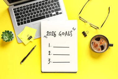 Listesi boş bir defter, dizüstü bilgisayar ve sarı arka planda bir fincan kahve. Yeni yıl hedefleri