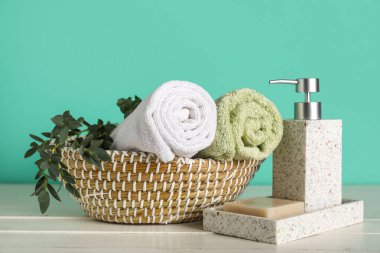 Temiz yumuşak havlular, hasır kase, sabun çubuğu ve turkuaz duvarın yanındaki beyaz masada bir şişe sıvı sabun, yakın plan.