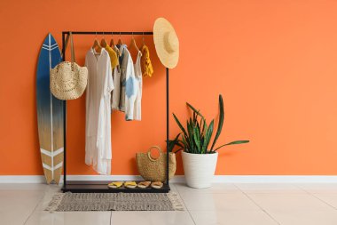 Sörf tahtası, ev bitkisi ve siyah askılık. Odada turuncu duvarın yanında şık kıyafetler var.