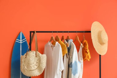 Sörf tahtası ve siyah askılık, turuncu duvarın yanında şık kıyafetler, yakın plan.