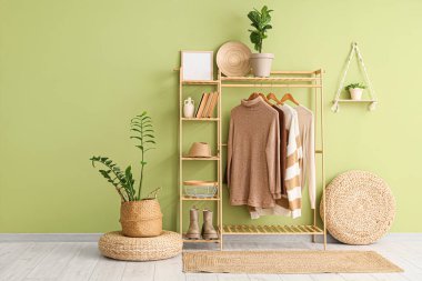 Poufs, ev bitkileri ve ahşap askılar. Odanın içinde yeşil duvarın yanında şık kıyafetler var.