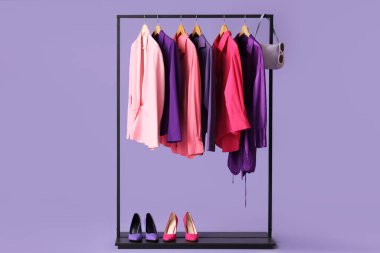 Mor arka planda şık kıyafetler, ayakkabılar ve aksesuarlar olan siyah askılar.