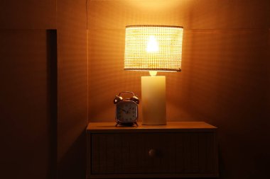 Kahverengi duvarın yanında parlayan lamba ve alarm saati olan komodinin yanında.