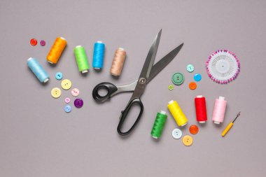 Gri arkaplanda makas, düğme ve dikiş iğneleri bulunan farklı iplik makaraları