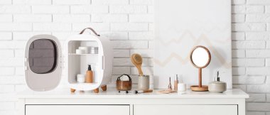 Cilt ürünleri olan küçük bir kozmetik buzdolabı, odadaki beyaz tuğla duvarın yanındaki çekmecelerin üzerindeki ayna ve resim. Yakın çekim.