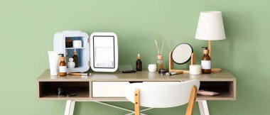 Küçük kozmetik buzdolabı, ayna ve lamba olan masa yeşil duvarın yanında.