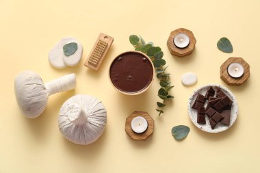 Yanan mumlar, bitkisel masaj torbası ve soluk sarı arka planda erimiş çikolatalı spa kompozisyonu.