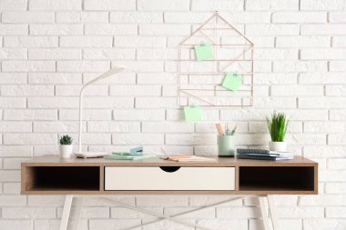 Masa lambası, ev bitkileri ve beyaz tuğla duvarın yanında ofis malzemeleri olan modern işyeri.