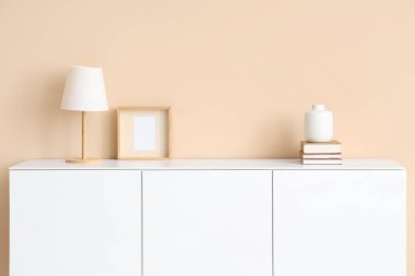 Masa lambası, fotoğraf çerçevesi, vazo ve odada bej duvarın yanındaki çekmecelerin üzerindeki kitaplar.