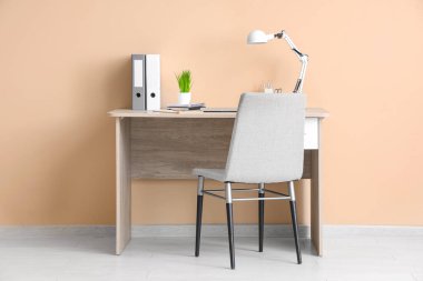 Masa lambası, ev bitkisi, ofis malzemeleri ve odada bej duvarın yanındaki sandalye olan modern işyeri.