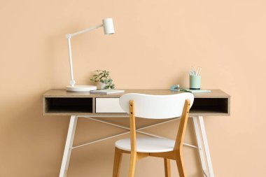 Masa lambası, ev bitkisi, ofis malzemeleri ve odada bej duvarın yanındaki sandalye olan modern işyeri.