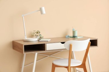 Masa lambası, ev bitkisi, ofis malzemeleri ve odada bej duvarın yanındaki sandalye olan modern işyeri.