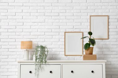 Odadaki beyaz tuğla duvarın yanındaki çekmecelerin üzerindeki lamba ve bitkiler, yakın plan.