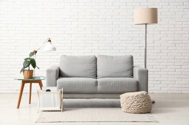 Oturma odasının içindeki beyaz tuğla duvarın yanında kanepe, lamba ve puf.
