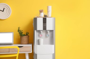 Kağıt bardaklı modern su soğutucusu ve odada sarı duvarın yanında dizüstü bilgisayarı olan iş yeri.