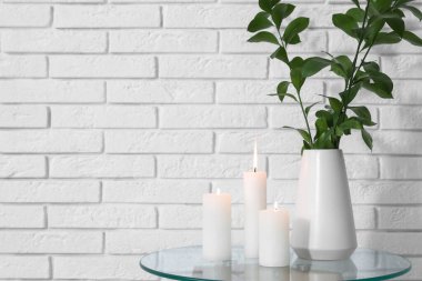 Odadaki beyaz tuğla duvarın yanındaki masada, vazoda yanan mumlar ve yeşil dallar.
