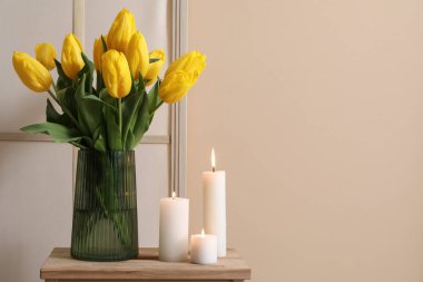Oturma odasındaki bej duvarın yanında vazoda mumlar ve çiçekler yanıyor.