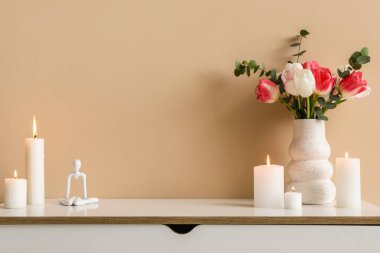 Yanan mumlar, heykelcikler ve vazolar. Odadaki bej duvarın yanındaki çekmecelerde çiçekler, yakın plan.