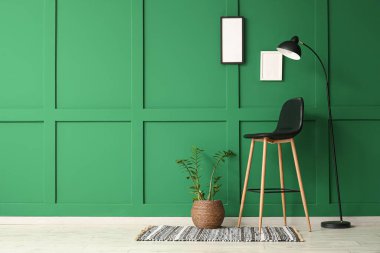 Yeşil duvarın yanında sandalye, çiçek ve zemin lambası olan modern tarz bir oda.