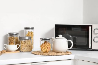 Mutfaktaki masada beyaz çaydanlık, kavanoz ve mikrodalga fırın, yakın plan.