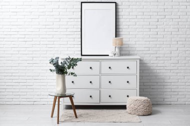 Beyaz tuğla duvarın yanında okaliptüs dallı vazo ve çekmeceli odanın içi.