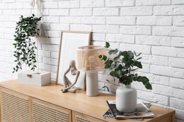 Fotoğraf çerçevesi, lamba ve vazo. Okaliptüs dalları, beyaz tuğla duvarın yanındaki çekmecelerde.