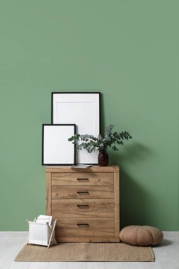 Blank photo frames and vase with eucalyptus twigs on chest of drawers near green wall in room