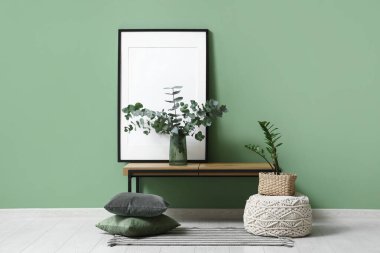 Blank photo frame and vase with eucalyptus twigs on bench near green wall in room