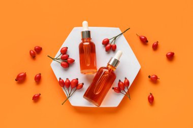 Decorative podium with bottles of cosmetic rose hip oil and berries on orange background