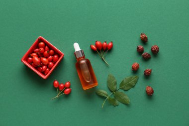 Bottle of cosmetic rose hip oil and bowl with berries on green background