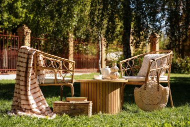 Sepeti, battaniyesi, çaydanlığı ve vazosu olan rahat hasır koltuklar parkta, açık havada kahve masasında.