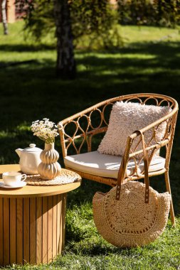 Rahat hasır koltuk şık bir çanta, çaydanlık ve bir vazo papatya çiçeği parktaki kahve masasında, açık havada