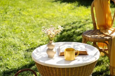 Rahat hasır koltuk, kitap, çay fincanı ve bir vazo papatya çiçeği parktaki sehpada, açık havada.